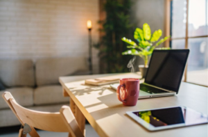 A desk with a laptop, red mug, notebook, and tablet. Sunlight illuminates the space. Sofa and potted plant in the background.